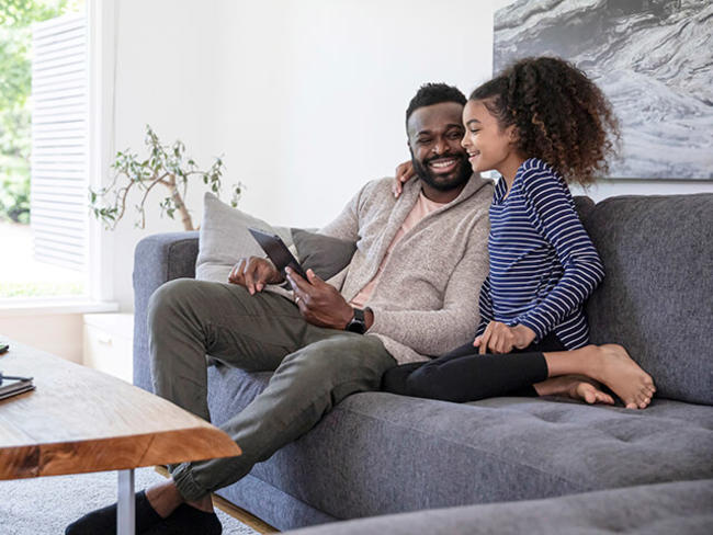 A father and daughter sit in living room reviewing information on tablet device